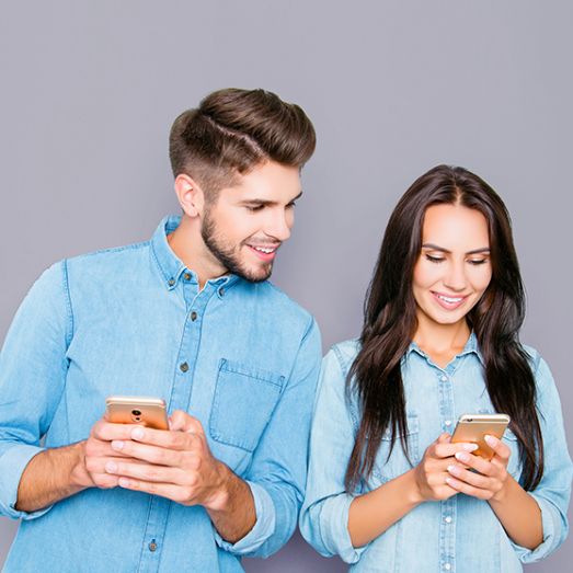 A man and woman, both with dark hair, stand closely together. The man wears a light blue shirt, while the woman has a cream-colored outfit. They stand against a light gray background.